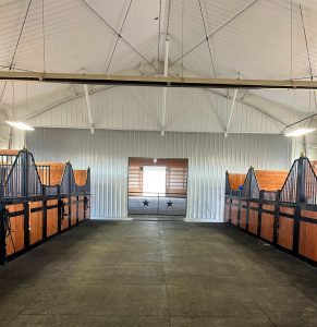 A clean, empty horse stable with wooden stalls on both sides and a window with two star decorations at the far end.