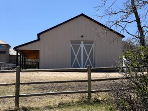 A beige barn with large white double doors sits behind a wooden fence on a sunny day; there is a covered area on the left side and sparse vegetation in the foreground.