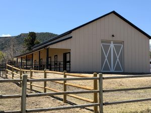 A tan barn with a large white sliding door and black trim is surrounded by a wooden fence, set in a rural area with hills and blue sky in the background.