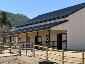 A beige barn with a black metal roof and several doors stands behind a wooden and metal fence on a sunny day, with trees and hills in the background.