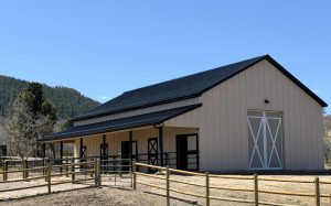 A beige barn with a black roof, large white double doors, and multiple stalls, surrounded by a wooden fence under a clear blue sky.