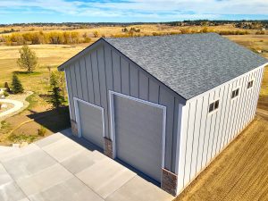 A large gray metal garage with two roll-up doors, situated on a concrete driveway in a rural area with open fields and trees.