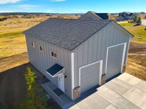 A tall, gray metal garage building with two large roll-up doors and a pedestrian entrance, situated on a paved driveway in a rural landscape.