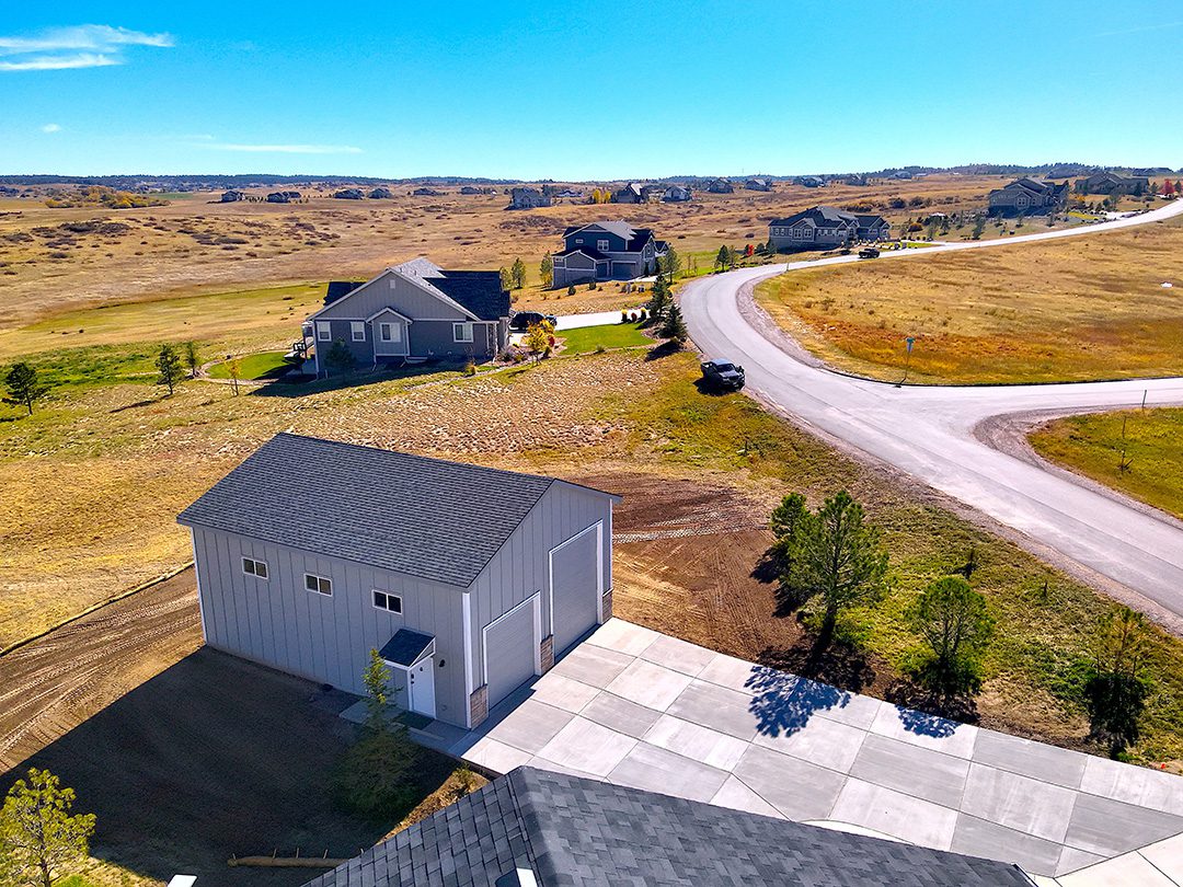 A large detached garage or workshop with a paved driveway is situated near a suburban home, complemented by a custom horse barn nearby, with open fields and a winding road in the background under a clear sky.