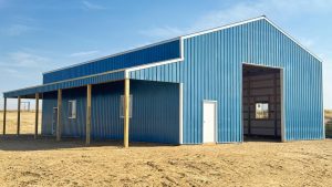 A large blue metal barn with a covered porch and several doors, situated on bare ground under a clear sky.