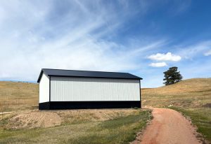 A black and white metal storage building sits on a dirt lot next to a winding gravel road, with grassy hills and a partly cloudy sky in the background.