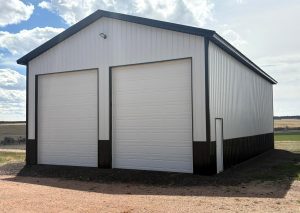 A large white metal storage building with two tall garage doors and a small side door, set on gravel with open land and sky in the background.