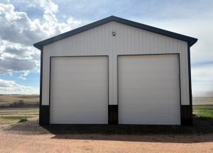 A two-door, white metal garage with dark trim stands on a gravel driveway in a rural landscape under a partly cloudy sky.
