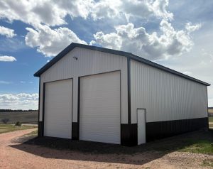 A large metal building with two tall garage doors and a smaller entry door stands on a gravel driveway under a partly cloudy sky.