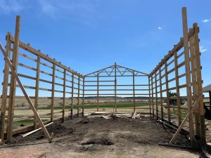 A wooden framework of a large building under construction stands on dirt ground with open fields in the background.