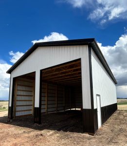 A large, partially open-sided metal barn with a high roof stands on bare ground under a blue sky with clouds.