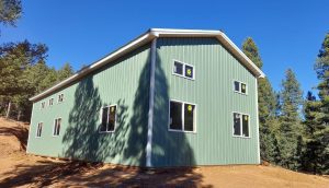 A green metal-sided building with multiple windows stands on a dirt clearing, surrounded by trees under a clear blue sky.