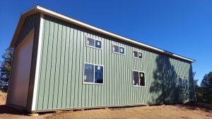 A large green metal building with multiple windows and a white garage door stands on a dirt lot under a clear blue sky.