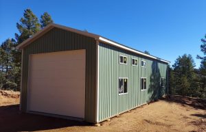 A green metal building with a large white garage door and several windows, situated on bare ground surrounded by trees under a clear blue sky.