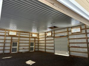 Interior of a large, unfinished garage or storage building with exposed wooden framing, metal ceiling, gravel floor, several small windows, two doors, and one roll-up garage door.