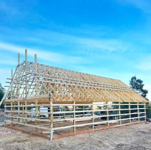 A wooden frame structure of a building under construction stands on a dirt lot, with a clear blue sky in the background.