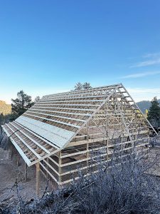 A large wooden barn frame under construction stands on dirt ground with trees and hills in the background under a clear blue sky.