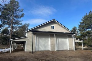 A large light gray detached garage with two tall white roll-up doors, a black roof, and a covered side area, surrounded by trees and dirt ground.
