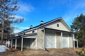 A large beige barn with white trim, multiple garage doors, small windows, and cupolas, surrounded by trees under a blue sky.