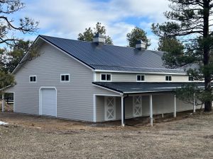 A large light gray barn with a black metal roof, multiple windows, and two cupolas, surrounded by trees and dirt ground.