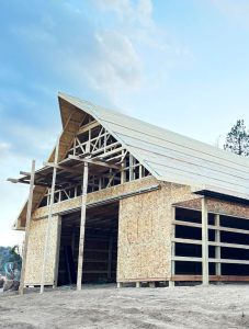 A large wooden barn under construction, featuring exposed framing and unfinished walls, set against a blue sky.