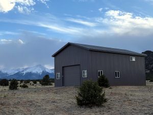 A gray metal building with a large garage door and several windows stands in a dry field with sparse bushes, mountains, and cloudy sky in the background.