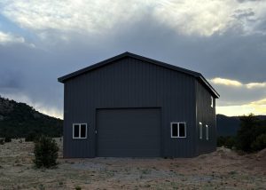 A large, dark gray metal building with a central garage door and four windows stands on a dirt lot with mountains and cloudy sky in the background.
