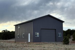 A gray metal building with a large garage door and several windows sits on a dirt lot with sparse vegetation under a cloudy sky.
