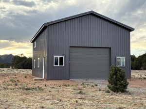 A gray metal building with a large garage door and two windows stands on a dry, grassy lot with trees and mountains in the background.
