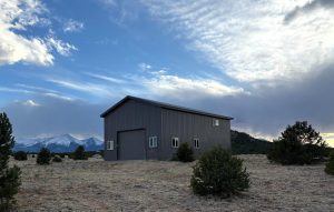 A large gray metal building with multiple windows and a garage door stands in an open, grassy field with distant mountains and trees under a partly cloudy sky.