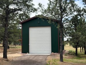 A green metal storage building with a large white garage door stands among tall pine trees on a dirt lot.