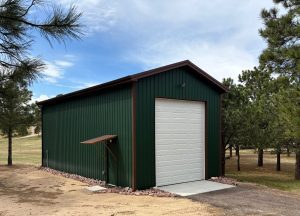 A green metal storage building with a large white garage door and a small awning, surrounded by trees on a dirt and grass lot.