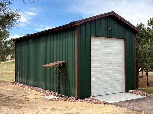 A green metal storage building with a large white garage door and a small side entrance, surrounded by trees and rocks.