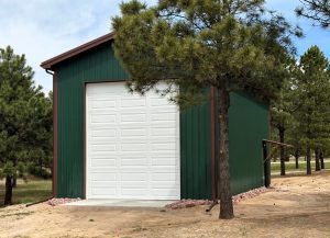 A green metal garage with a large white roll-up door stands among pine trees on a dirt lot.