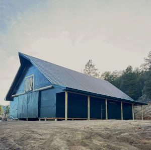 A large, unfinished barn-like building covered in dark plastic sheeting stands on a dirt lot, surrounded by trees and a cloudy sky.