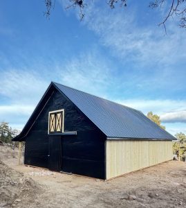 A large barn with a black exterior and light wood siding stands on a dirt lot under a blue sky with scattered clouds.