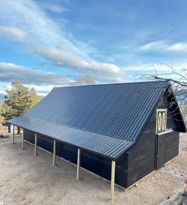A black, barn-style structure with a sloped metal roof and wooden framing stands on bare ground under a partly cloudy sky.