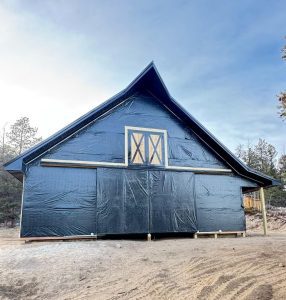 A large, partially constructed building is wrapped in a dark weather-resistant material, set on a dirt lot with trees in the background under a clear sky.