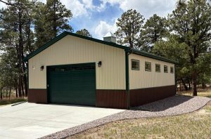 A beige and brown detached garage with a green door sits on a concrete driveway, surrounded by trees and gravel landscaping.