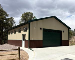A beige and brown metal garage with a large green door, small windows, and outdoor lights, set on a concrete driveway beside trees under a cloudy sky.