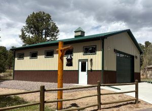 A tan and brown metal outbuilding with a green roof stands behind a wooden fence, surrounded by trees; a wooden post with a lantern is in the foreground.