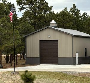 A metal garage building with a large roll-up door sits next to a concrete driveway; an American flag is on a pole nearby, surrounded by pine trees.