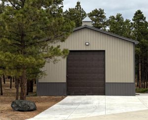 A gray metal garage with a closed dark brown door sits beside pine trees on a concrete driveway in a wooded area.