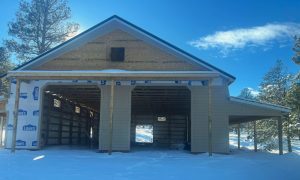 A partially constructed garage with exposed framing and siding, surrounded by snow and trees, under a clear blue sky.