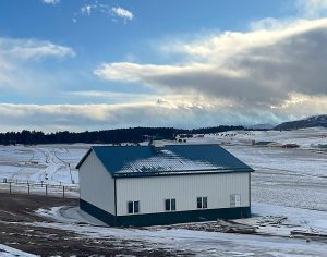 A green-roofed white barn sits on a snowy field under a partly cloudy sky, with tire tracks visible in the snow and distant hills in the background.
