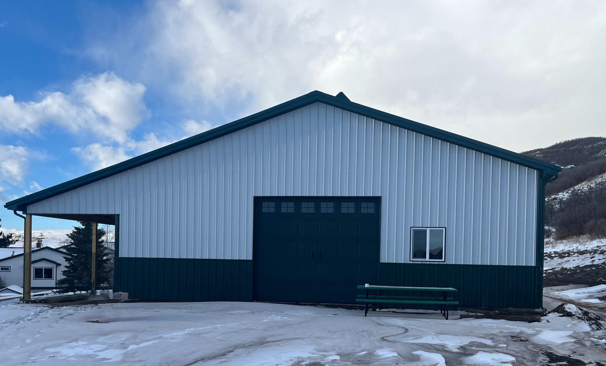 A large metal building with a green roof and trim, a wide garage door, a single window, and a bench in front, surrounded by snow and cloudy skies.