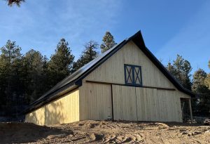 A newly constructed light wood barn with a metal roof stands on dirt ground, surrounded by pine trees under a clear blue sky.