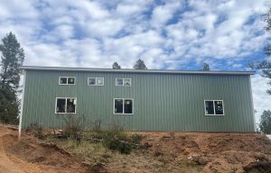 A green metal-sided building with several windows sits on a dirt slope under a partly cloudy sky, surrounded by sparse trees and brush.