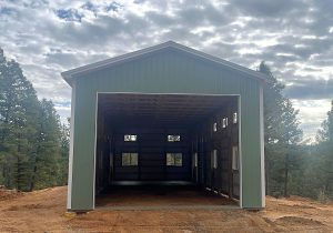 A large, unfinished metal building with an open front, set on a dirt lot surrounded by trees under a cloudy sky.