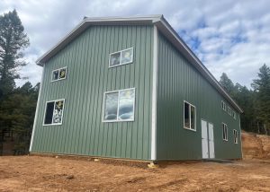 A green metal building with white trim and several windows, situated on bare ground with trees and a cloudy sky in the background.